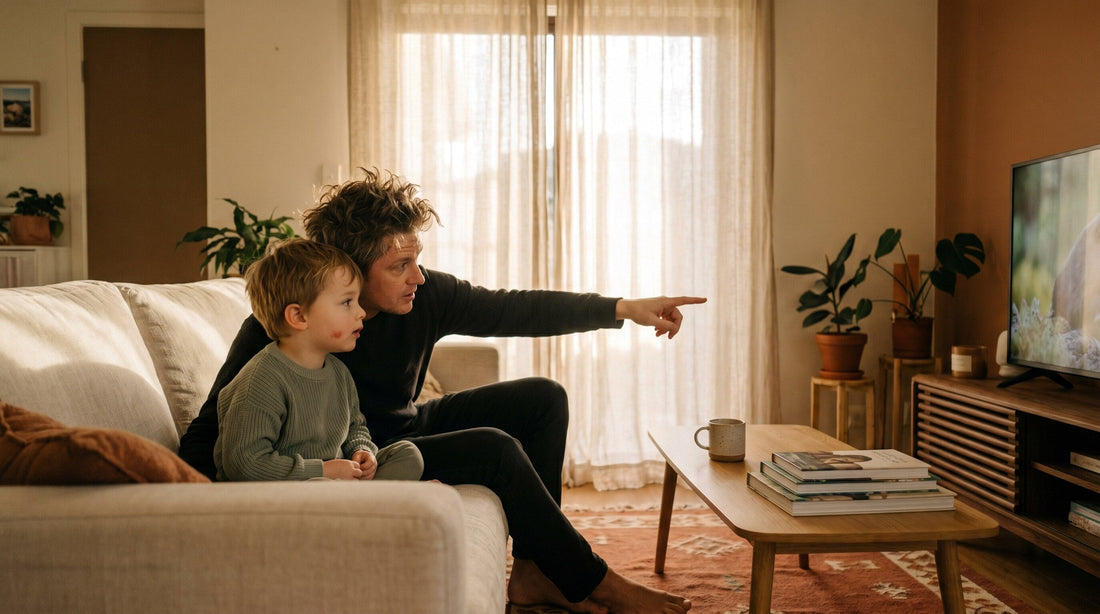 A child sitting on a couch with a parent, both watching a screen together, the parent pointing at something and the child looking curious rather than passive. Warm afternoon light in a home living room.