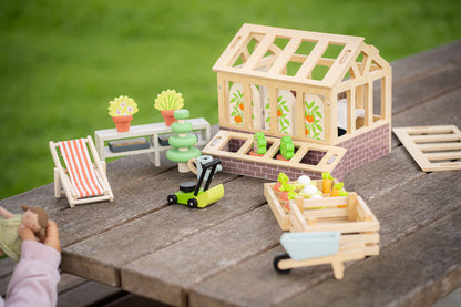 Miniature garden scene with a wooden greenhouse, plants, and small toys on a wooden surface.