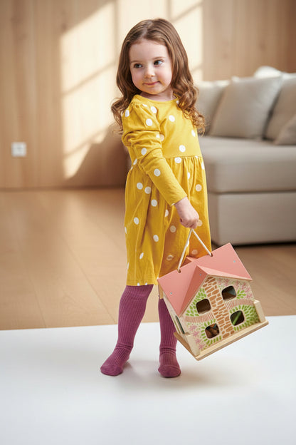 Young girl in a yellow dress with white polka dots holding a pink toy house against a light gray background